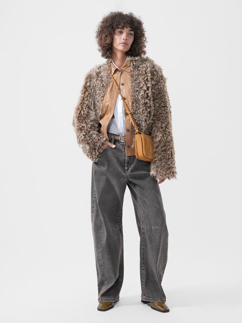 Une personne portant une veste à texture duveteuse sur une chemise marron clair à col blanc, un jean gris ample et un sac à bandoulière marron clair.