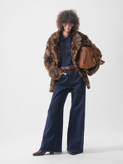 Une personne portant un manteau de fourrure marron sur une combinaison en jean foncé avec une ceinture marron et tenant un sac en daim marron.