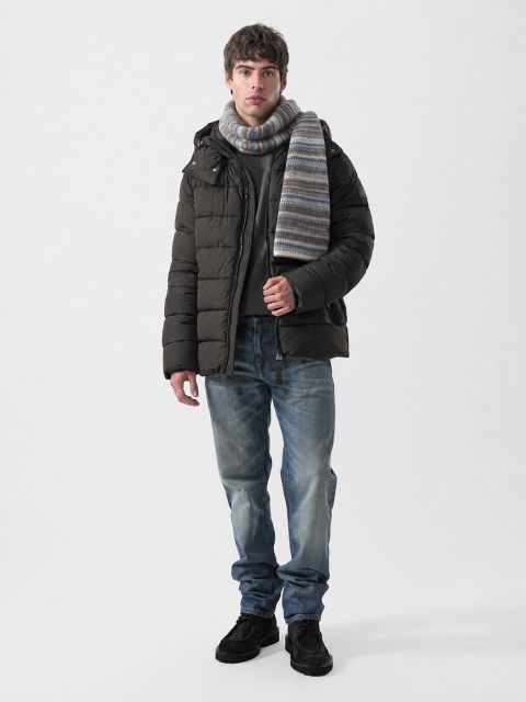 Un homme portant une doudoune marron foncé, un pull gris, une écharpe rayée, un jean bleu et des chaussures foncées.