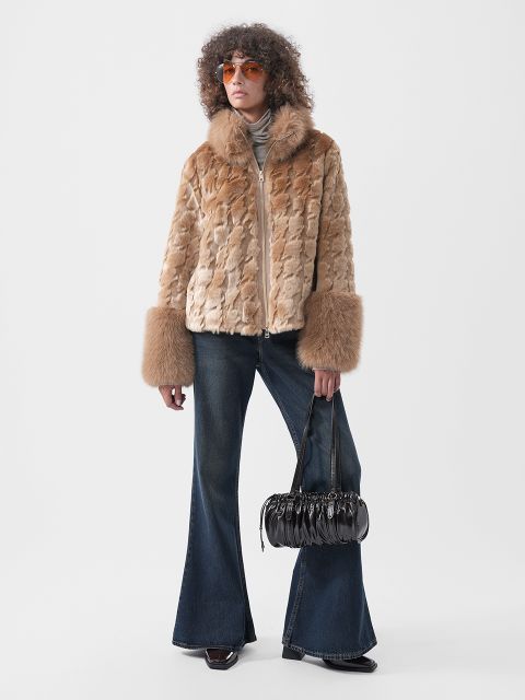 Une personne portant une veste en fausse fourrure marron clair avec de larges poignets, un jean évasé foncé et des lunettes de soleil orange, tenant un sac à main plissé noir.