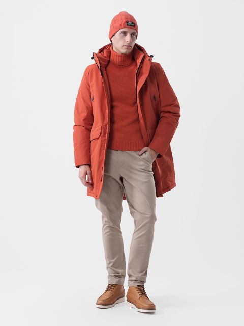 Un homme portant une casquette orange, un col roulé et une parka, ainsi qu'un pantalon beige et des bottes en cuir marron.