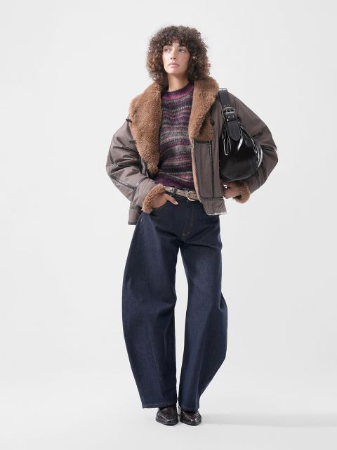 Une personne portant une veste marron avec un col en fourrure, un pull rayé violet et un jean foncé à jambe large, et portant un sac à bandoulière noir.