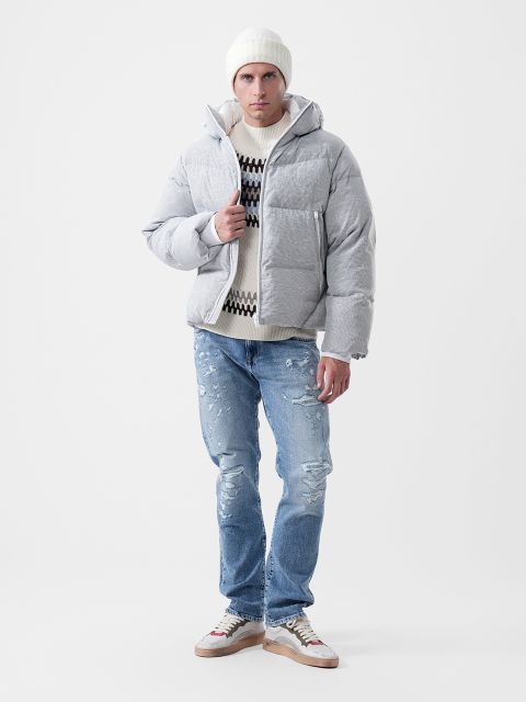 Un homme portant une doudoune gris clair, un pull à motifs, un bonnet blanc, un jean bleu délavé et des baskets.