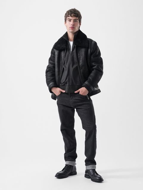 Un homme portant un blouson aviateur noir doublé de peau de mouton, un jean noir et des bottes noires.