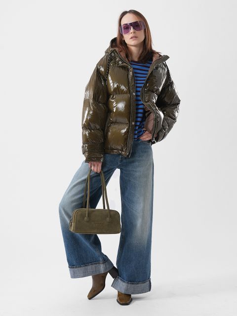 Une personne portant une doudoune vert olive brillante, une chemise à rayures bleues, un jean large et des lunettes de soleil violettes, tenant un sac à main vert olive.