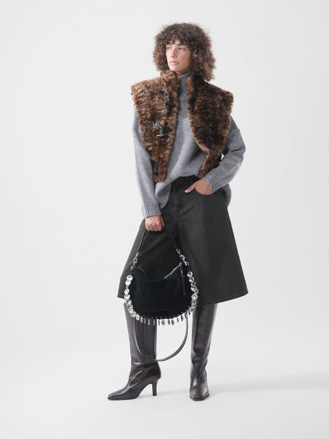 Un mannequin porte un pull à col roulé gris, un gilet en fausse fourrure marron et une jupe en jean noire ; elle tient un sac à main noir avec des ferrures rondes argentées et porte de hautes bottes marron.