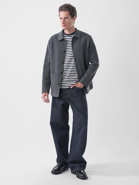 Un homme portant une veste grise sur un t-shirt rayé bleu marine et blanc, un jean bleu foncé et des mocassins noirs.