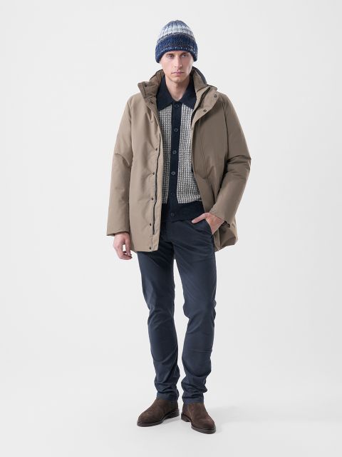 Un homme portant un parka beige clair sur un pull à motifs, un pantalon foncé, des bottes marron et un bonnet tricoté rayé.