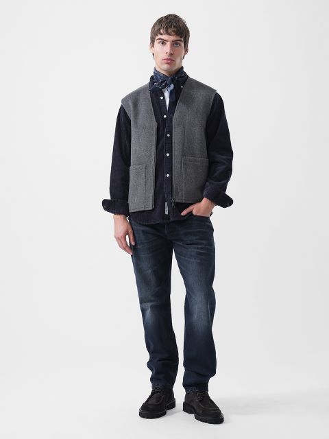 Un homme portant un gilet gris sur une chemise boutonnée foncée, un foulard à motifs, un jean bleu foncé et des bottes foncées.